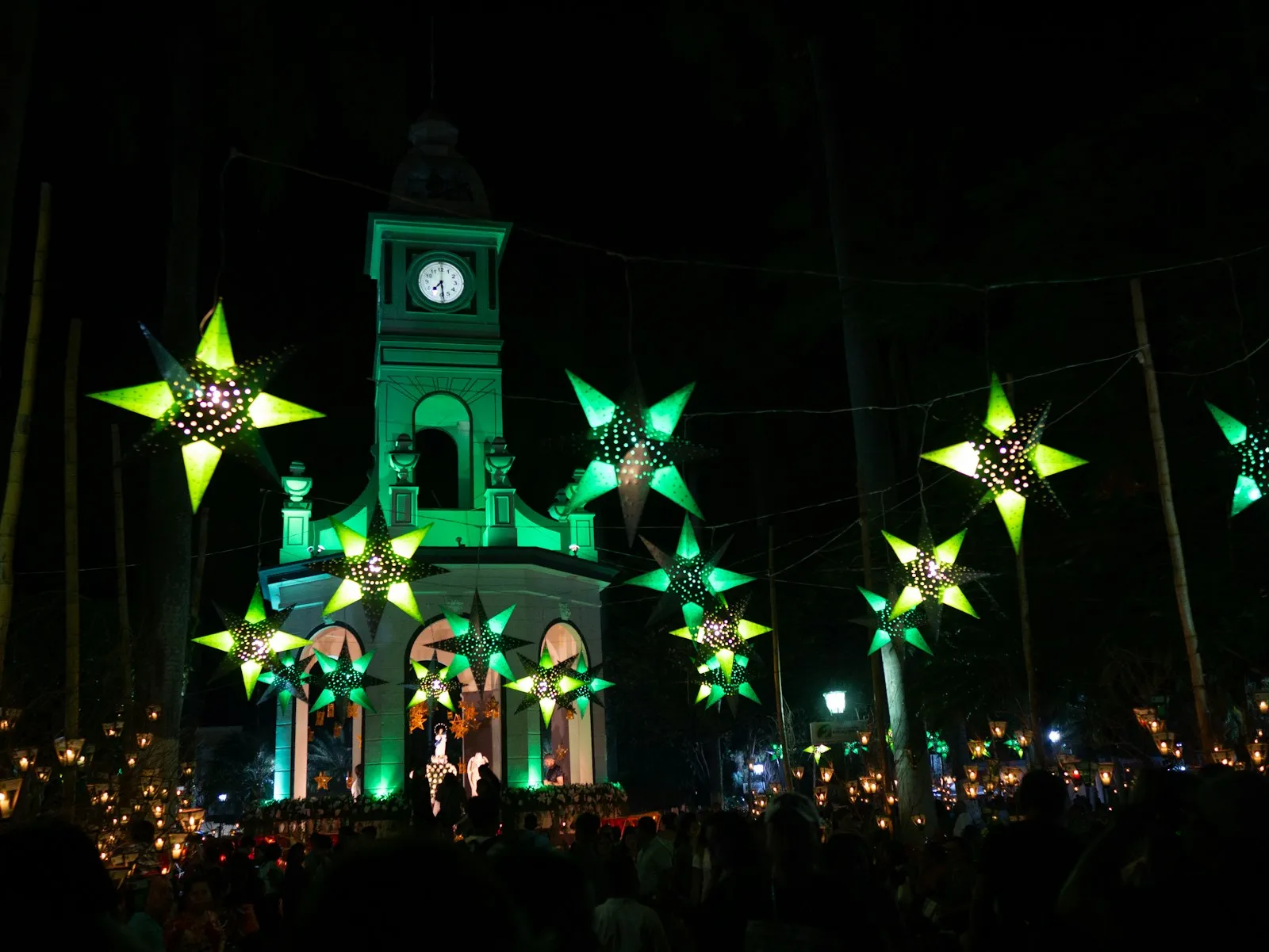 Torre del reloj iluminada de verde durante la noche del Día de los Farolitos en Ahuachapán, decorada con estrellas luminosas y rodeada de farolitos encendidos y público