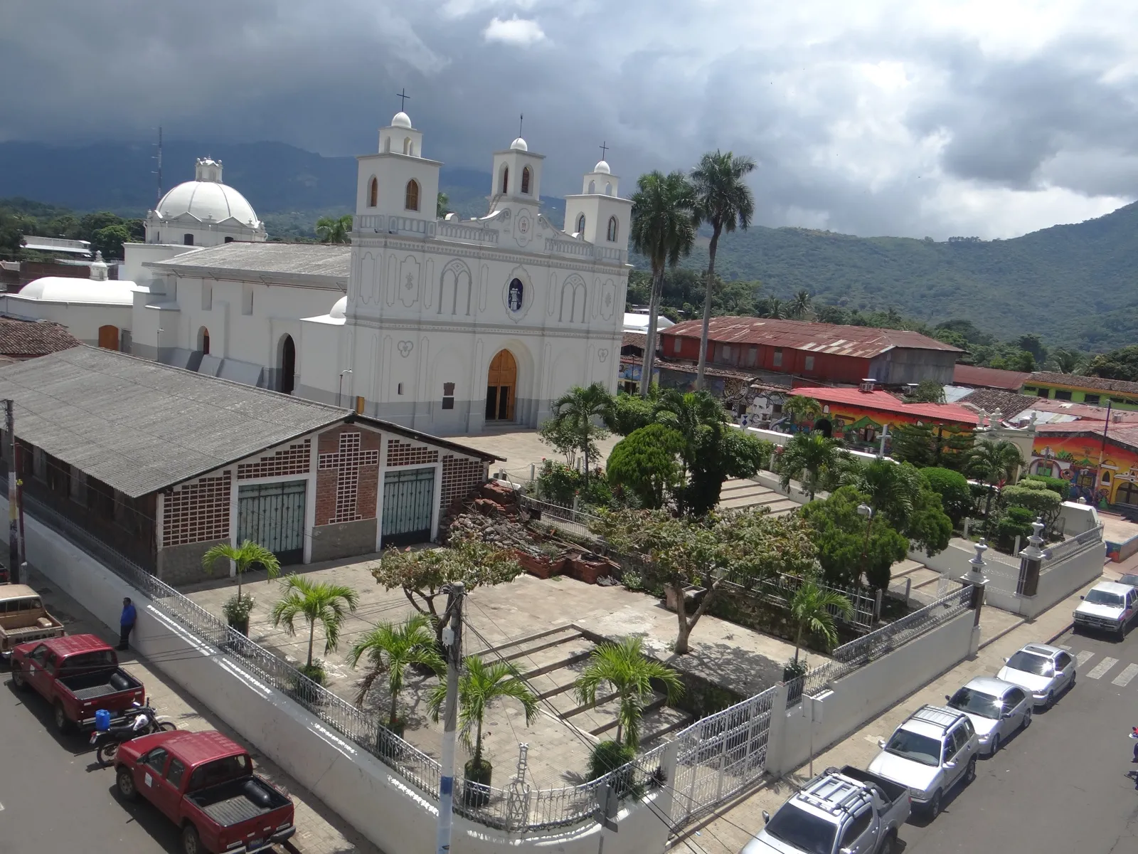 Vista elevada de la parroquia Nuestra Señora de la Asunción de Ahuachapán, iglesia blanca con torres y cúpula, rodeada de árboles y montañas