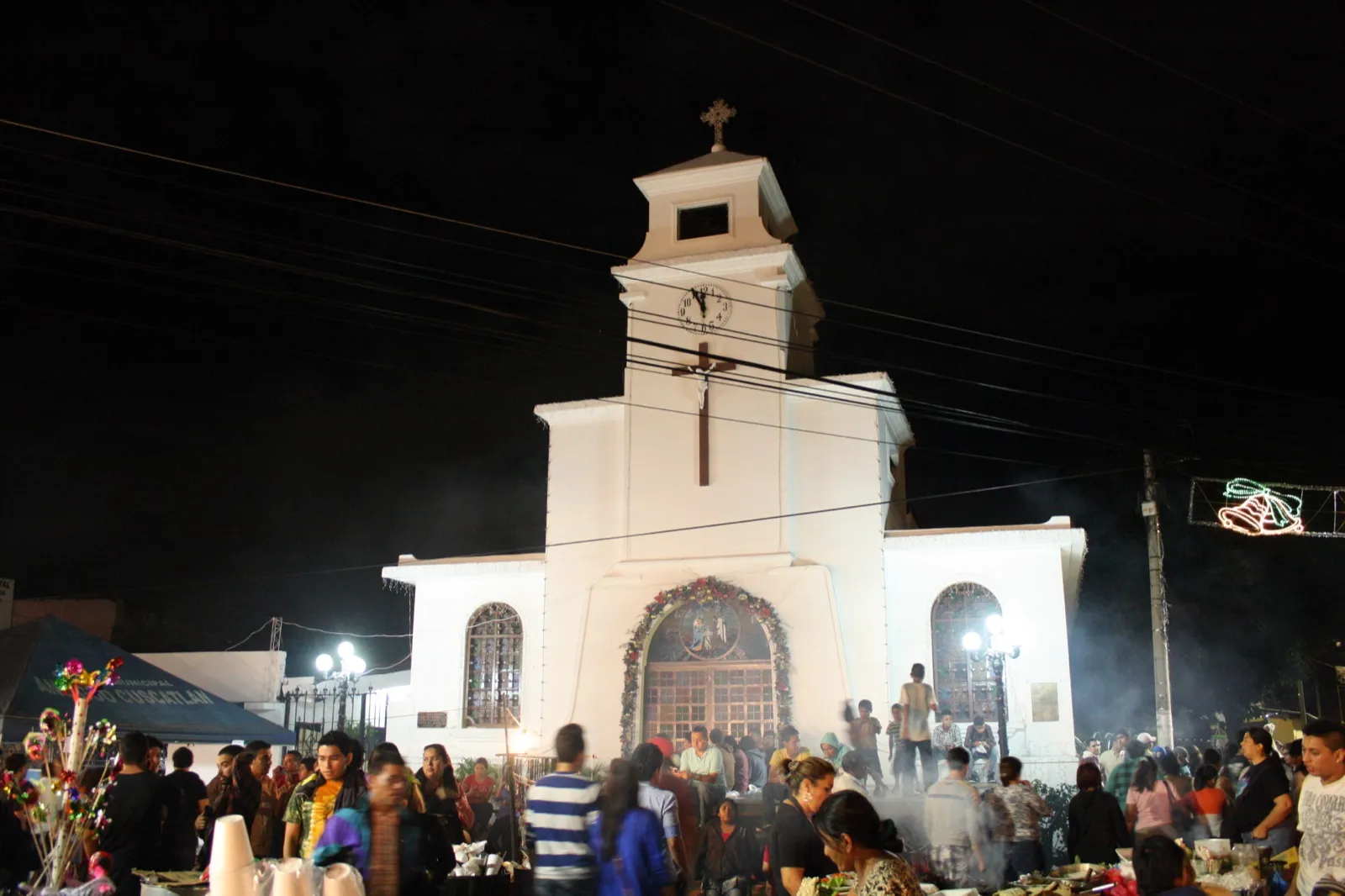 Iglesia de los Santos Niños Inocentes de Antiguo Cuscatlán iluminada de noche durante sus fiestas patronales, con público en la plaza